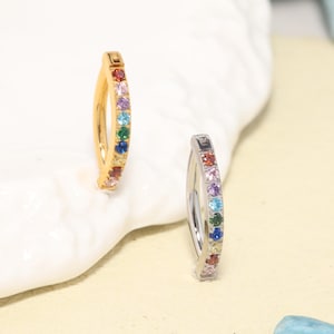 May include: Two curved navel rings, one gold-toned and one silver-toned, each featuring a vertical row of seven small, multi-colored round gemstones including red, pink, purple, light blue, green, dark blue, and yellow stones set in metal.