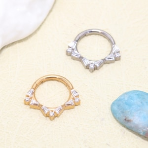 Peut inclure: Deux anneaux circulaires à charnière, l'un argenté et l'autre doré, chacun doté d'un arc décoratif orné de petites pierres transparentes taillées en baguette et en rond le long du bord inférieur, créant un design symétrique et étagé.
