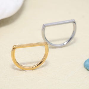 May include: Two D-shaped rings, one in a polished gold tone and one in a polished silver tone. Each ring features a straight horizontal bar across the top and a curved semi-circular bottom section, constructed from smooth, solid metal material.