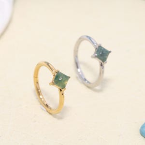 May include: Two rings featuring a square-cut green stone set in metal. One ring is gold-toned, and the other is silver-toned. Each ring has a thin, circular band and a prong setting that secures the square gemstone at the top of the band.