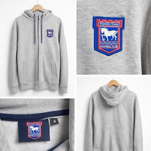May include: A gray zip-up hoodie with a blue and red Ipswich Town Football Club patch on the chest. The patch features a white horse and the words "IPSWICH TOWN FOOTBALL CLUB". The hoodie has a drawstring hood and a front pocket.