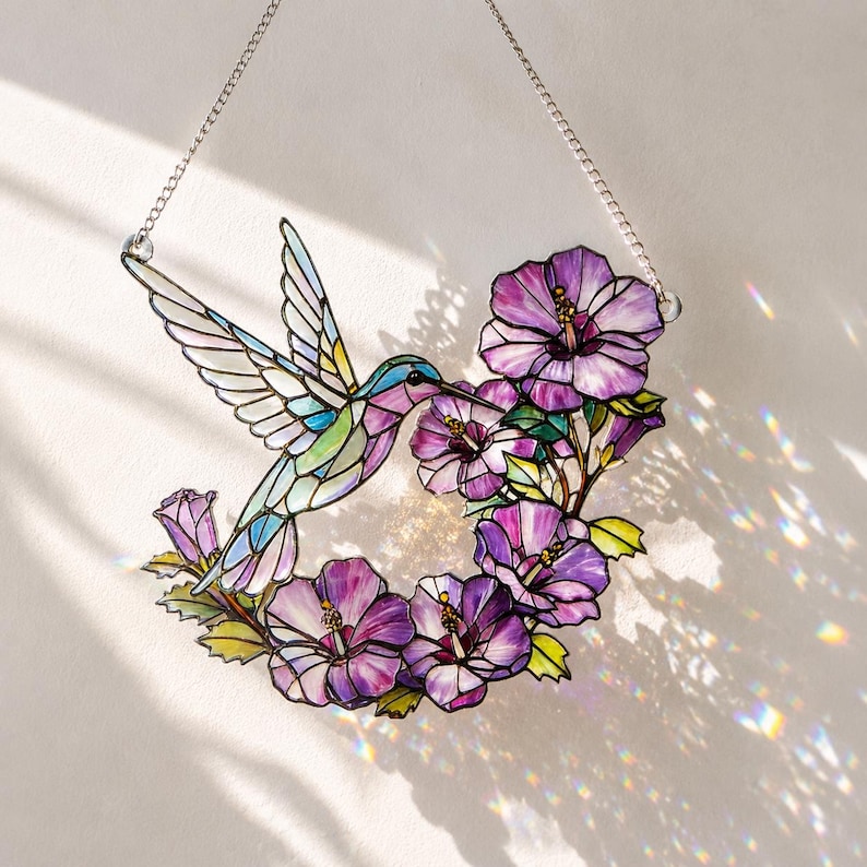 May include: A stained glass hummingbird with outstretched wings, feeding from purple hibiscus flowers. The colorful bird features shades of blue, green, and pink. A silver chain is attached for hanging. The artwork is set against a white background.