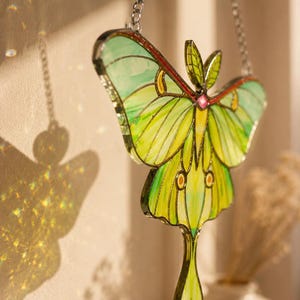 May include: A stained glass suncatcher shaped like a luna moth with light green and yellow wings, dark green veins, and a small pink accent at the center. The moth is suspended by a silver metal chain and features intricate soldered metal borders.