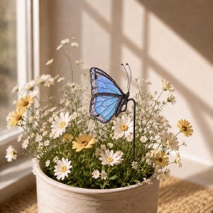 May include: A stained glass butterfly ornament in shades of light and dark blue with black outlines, mounted on a thin black metal stake. The butterfly features delicate wire antennae and is positioned among a cluster of white and yellow daisies.