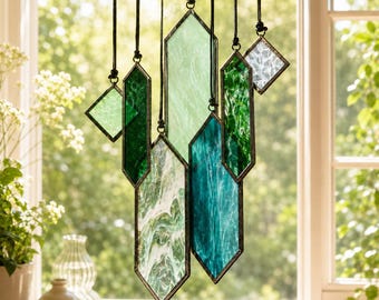 May include: A hanging mobile featuring multiple geometric stained glass pieces in various shades of green, teal, and white. The glass panels are framed in dark metal and suspended by black cords at different heights, creating a layered arrangement.