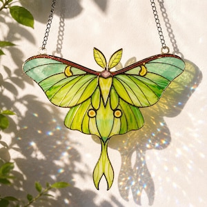 May include: A stained glass suncatcher shaped like a Luna moth with translucent lime green and pale yellow wings, dark metal soldering, and a small pink accent at the center. The moth hangs from a silver-toned metal chain for display.