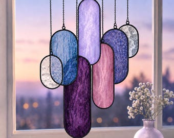 May include: A hanging suncatcher composed of multiple vertical oval-shaped stained glass panels in shades of purple, blue, pink, and clear textured glass. Each panel is framed in black metal and suspended by thin black chains at varying heights.