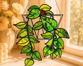 May include: A stained glass suncatcher featuring a trailing green plant with heart-shaped leaves and a small red ladybug with black spots. The plant is contained within a clear triangular glass frame and hangs from a silver metal chain.