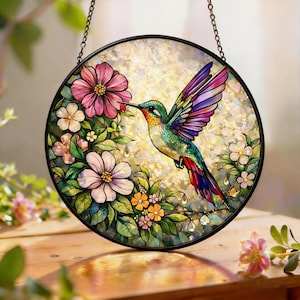 May include: A round stained glass window featuring a colorful hummingbird with outstretched wings, feeding from a pink flower. The bird is surrounded by other flowers and foliage, with a shimmering background and a black metal frame.