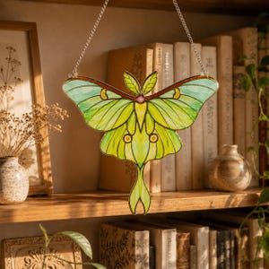 May include: A stained glass suncatcher shaped like a Luna moth with light green and yellow wings, dark green leaf-like upper wings, and a small pink center accent. The moth is suspended by a silver metal chain against a bookshelf background.