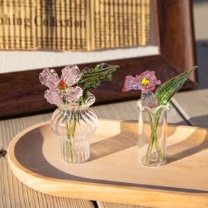 May include: Two glass flower arrangements sit on a wooden tray. One arrangement features a pink flower with a red center in a small, clear vase. The other has a purple flower with a yellow center in a clear, slender vase. Green glass leaves are included.