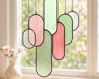 May include: A hanging window ornament featuring multiple elongated oval panels of textured stained glass in shades of pale green, soft pink, and translucent white, each framed in black metal and suspended by thin black chains from above.