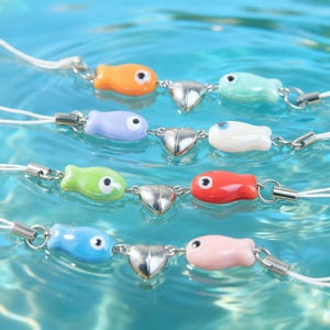 Puede incluir: Una colección de colgantes coloridos con forma de pez, cada uno conectado a un dije plateado en forma de corazón. Los colgantes son de varios colores, incluyendo naranja, verde, azul y rosa, y están ensartados en cordones blancos. El fondo es una superficie azul claro, similar al agua.