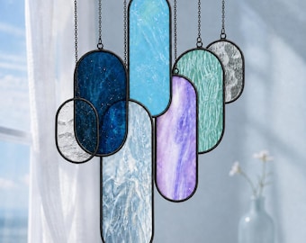 May include: A hanging mobile featuring multiple oval-shaped stained glass panels in shades of deep blue, light blue, purple, teal, and clear textured glass. Each panel is framed in black metal and suspended by thin black chains at varying heights.