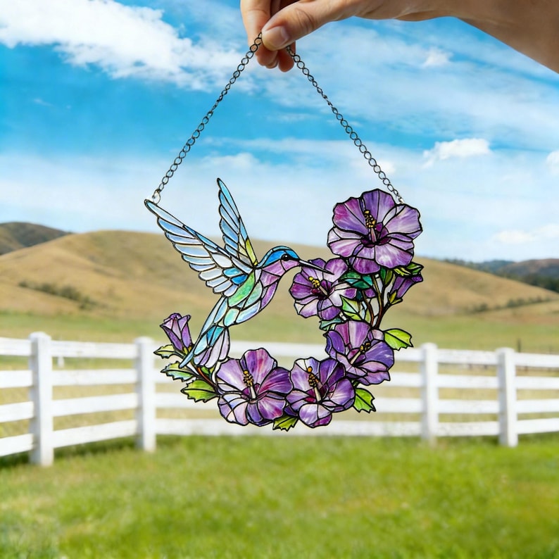 May include: Stained glass hummingbird suncatcher with a chain for hanging. The hummingbird is depicted with blue, green, and purple hues, hovering near a wreath of purple flowers. The background shows a green field, white fence, and blue sky.