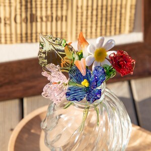 May include: A clear glass vase with a bouquet of colorful glass flowers. The flowers include a white daisy, red rose, blue flower, and pink flower. The vase sits on a wooden surface.