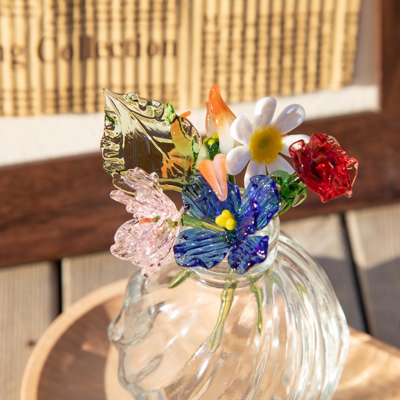 May include: A clear glass vase with a bouquet of colorful glass flowers. The flowers include a white daisy, red rose, blue flower, and pink flower. The vase sits on a wooden surface.