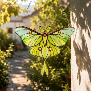May include: A stained glass suncatcher shaped like a luna moth with vibrant lime green and pale teal wings. The moth features dark outlines, a small pink center detail, and yellow crescent markings on the upper wings, suspended by a metal chain.
