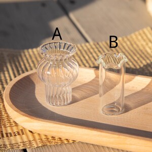 May include: Two small, clear glass vases on a wooden tray. Vase A has a bulbous body with vertical ridges and a flared rim. Vase B is a slender, cylindrical shape with a ruffled top. The background is blurred.