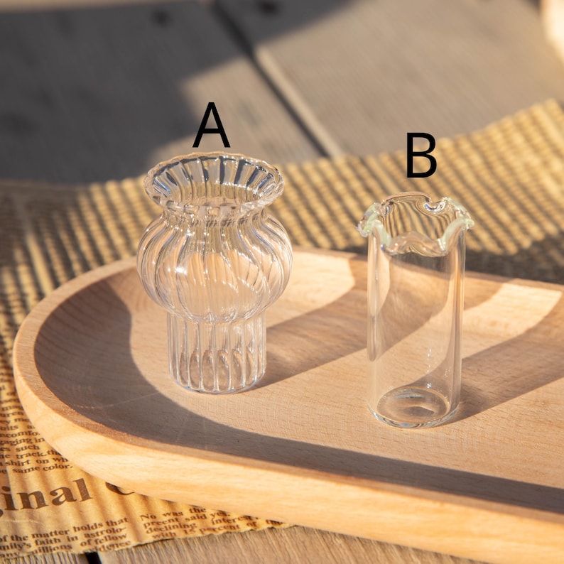 May include: Two small, clear glass vases on a wooden tray. Vase A has a bulbous body with vertical ridges and a flared rim. Vase B is a slender, cylindrical shape with a ruffled top. The background is blurred.