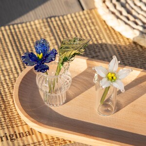 May include: Two glass flower sculptures in small vases on a wooden tray. One vase holds a blue flower with a yellow center and a green leaf. The other vase holds a white flower with a yellow center.