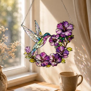 May include: Stained glass hummingbird with a colorful design, surrounded by purple hibiscus flowers. The artwork is suspended by a silver chain. The hummingbird has a long beak and outstretched wings, with a variety of colors including blue, green, and purple.
