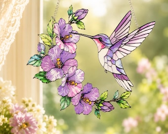 May include: A stained glass suncatcher featuring a hummingbird with purple and white wings hovering near a curved arrangement of purple hibiscus flowers and green leaves, suspended by a silver metal chain against a bright window.