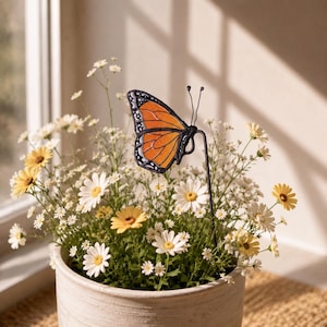 Può includere: Un ornamento a forma di farfalla monarca in vetro colorato su un sottile paletto di metallo nero, con ali arancioni, nere e bianche e delicate antenne in filo metallico, posizionato verticalmente in un vaso di margherite bianche e gialle.