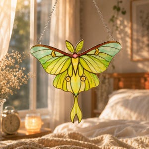 May include: A stained glass suncatcher shaped like a luna moth with vibrant lime green and pale yellow wings, featuring dark outlines, small circular accents, and a central pink diamond detail, suspended by a thin silver metal chain.