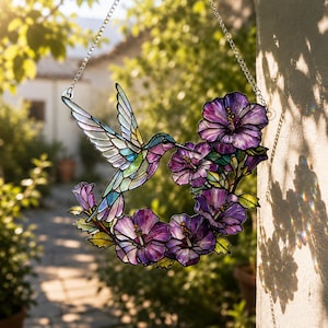 May include: Stained glass hummingbird suncatcher with a vibrant design. The bird is depicted with colorful wings and a long beak, hovering near a wreath of purple hibiscus flowers. It hangs from a silver chain, perfect for a window or garden decor.