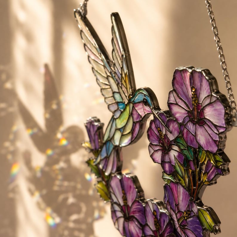 May include: Stained glass hummingbird with outstretched wings, feeding from purple hibiscus flowers. The colorful bird features shades of blue, green, and yellow. The flowers have purple petals and yellow centers. A silver chain is attached for hanging.