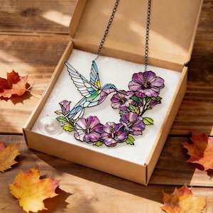 May include: A stained glass hummingbird and floral design necklace in a cardboard box. The hummingbird has blue, green, and white wings, with a purple and pink flower arrangement. The necklace hangs on a black chain. The box sits on a wooden surface with autumn leaves.