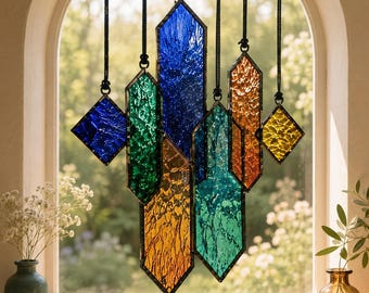 May include: A stained glass suncatcher featuring multiple geometric crystal-shaped panels in blue, green, orange, and yellow. Each textured glass piece is framed in dark metal and suspended by black cords, creating a vertical arrangement of colors.