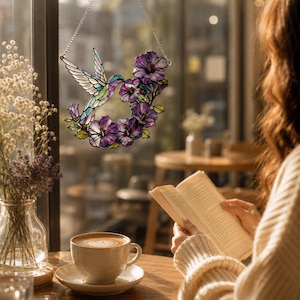 May include: A stained glass hummingbird suncatcher with a floral wreath of purple flowers. The hummingbird has iridescent wings and is suspended by a silver chain. The suncatcher is hanging in front of a window.