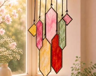 May include: A hanging mobile featuring multiple geometric stained glass pieces in shades of yellow, pink, red, and green. Each piece is framed in dark metal and suspended by thin black cords at varying heights, creating a layered, vertical arrangement.
