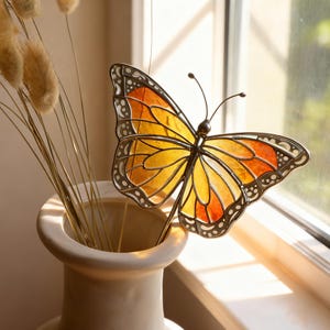 May include: A decorative butterfly stake with a stained glass design in shades of yellow, orange, and black. The butterfly is placed in a cream-colored vase with dried ornamental grass. The butterfly has a metal frame and is positioned near a window.
