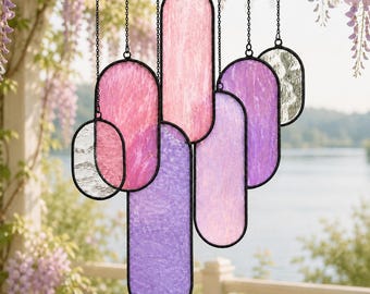 May include: A hanging suncatcher composed of multiple elongated oval glass panels in shades of pink, purple, and clear textured glass. Each panel is framed in a thin black metal border and suspended by individual black chains at varying heights.