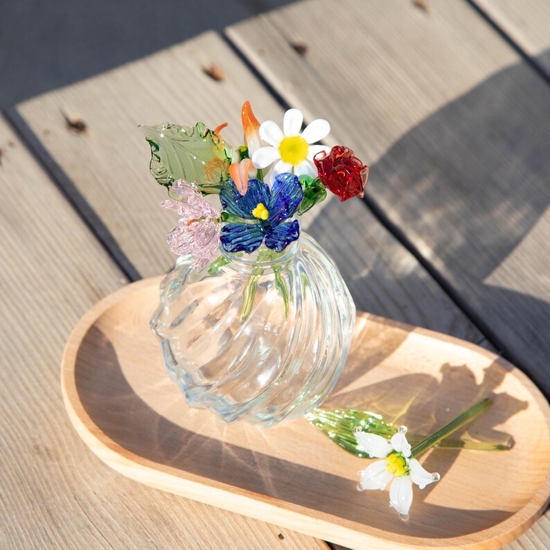 May include: A clear glass vase with a swirl design, holding colorful glass flowers. The flowers include a white daisy, red rose, blue pansy, and pink and orange accents. The vase sits on a wooden tray.