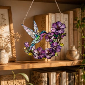 May include: A stained glass hummingbird with a colorful design, suspended by a silver chain. The bird is positioned near a wreath of purple flowers. The artwork is displayed on a wooden shelf with books and decorative items.
