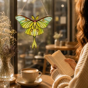May include: A stained glass suncatcher shaped like a luna moth with light green and yellow wings, dark outlines, and a small pink accent at the center. The moth is suspended by a silver-colored metal chain against a window pane.