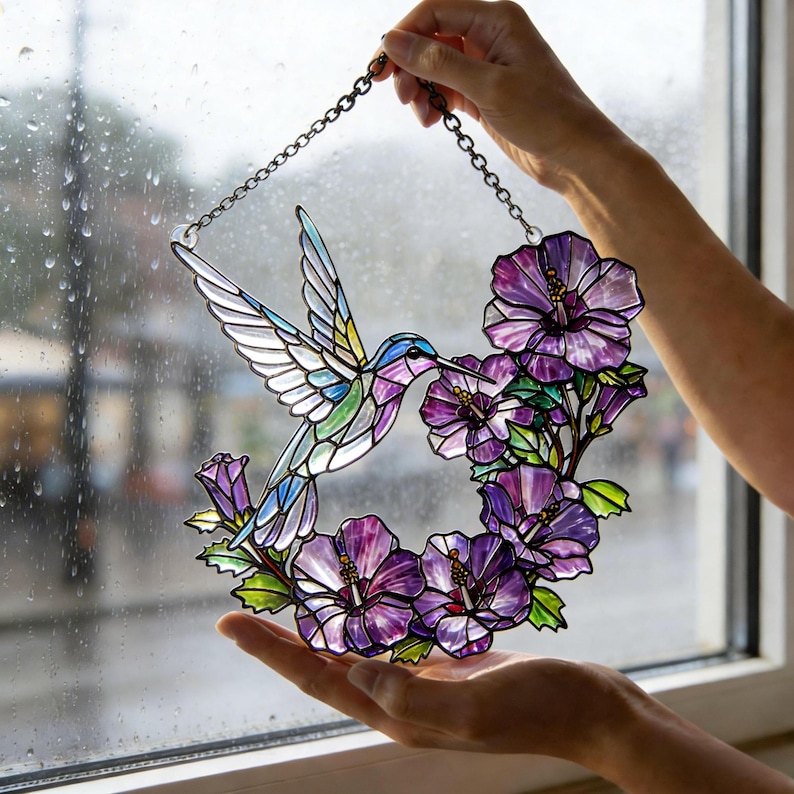 May include: Stained glass hummingbird and flower window hanging. The hummingbird is depicted with blue, green, and purple hues, with white wings. The flowers are purple and the leaves are green. The piece is held by a chain.
