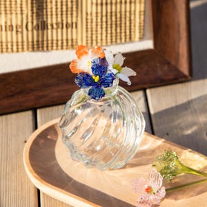 May include: A clear glass vase with a spiral design, holding colorful glass flowers in shades of blue, orange, and white. The vase sits on a wooden tray, with additional glass flowers nearby. The overall theme is decorative floral art.