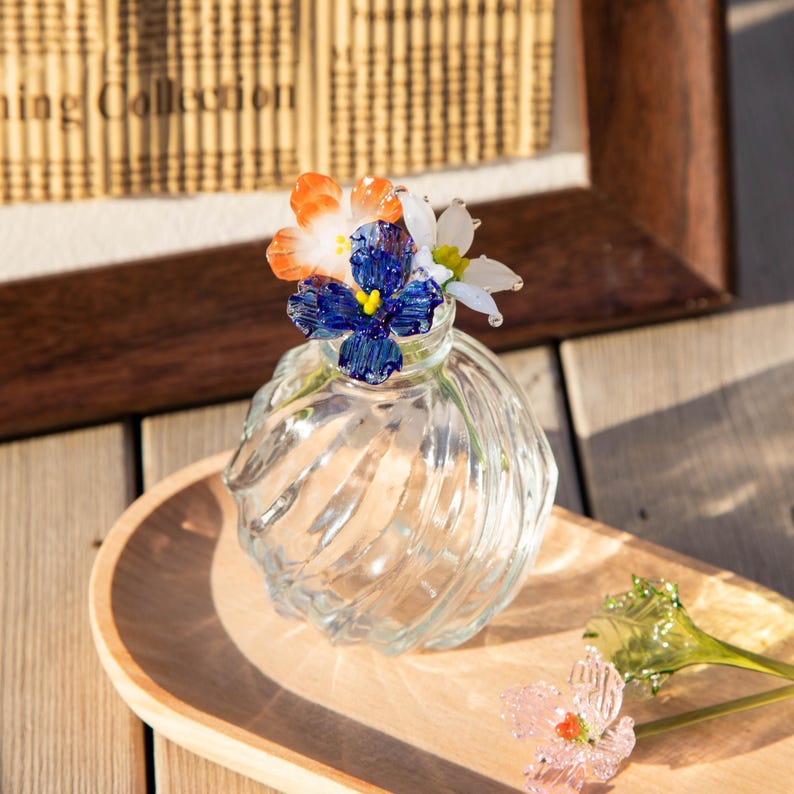 May include: A clear glass vase with a spiral design, holding colorful glass flowers in shades of blue, orange, and white. The vase sits on a wooden tray, with additional glass flowers nearby. The overall theme is decorative floral art.