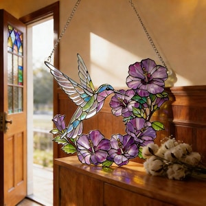 May include: Stained glass hummingbird with outstretched wings, feeding on purple hibiscus flowers. The colorful bird features shades of blue, green, and purple. The floral wreath is suspended by a silver chain, creating a decorative window or wall hanging.