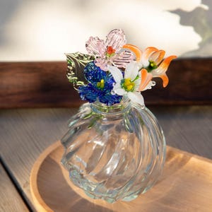 May include: A clear glass vase with a swirl design, topped with colorful glass flowers. The flowers include pink, blue, white, and orange petals with green leaves. The vase sits on a wooden tray.