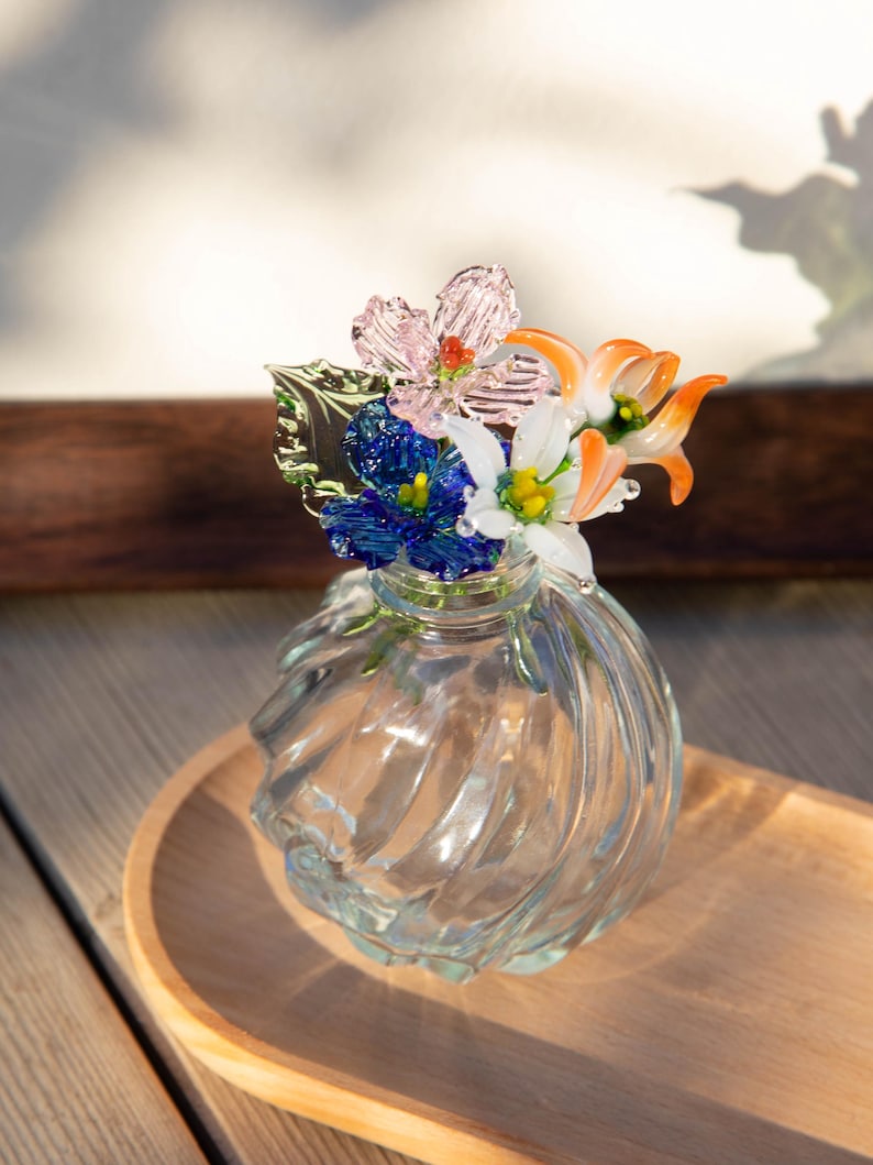 May include: A clear glass vase with a swirl design, topped with colorful glass flowers. The flowers include pink, blue, white, and orange petals with green leaves. The vase sits on a wooden tray.