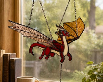 May include: A stained glass suncatcher shaped like a dragon with red and white body segments and textured amber wings. The dragon is suspended by thin metal chains, featuring dark leaded outlines that define its wings, tail, and body form.