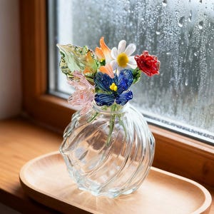 May include: A clear glass vase with a spiral design, holding colorful glass flowers. The flowers include orange, white, red, blue, and pink blooms, and green leaves. The vase sits on a wooden tray near a window.