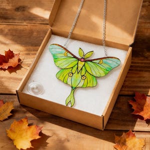 May include: A stained glass luna moth suncatcher with vibrant lime green and yellow wings, featuring dark brown outlines and a small pink center detail. The moth is attached to a silver metal chain and rests inside a cardboard box with white foam.