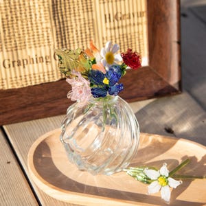 May include: A clear glass vase with a spiral design holds a colorful bouquet of glass flowers. The flowers include orange, blue, red, white, and pink blooms. A wooden tray and a framed document are in the background.